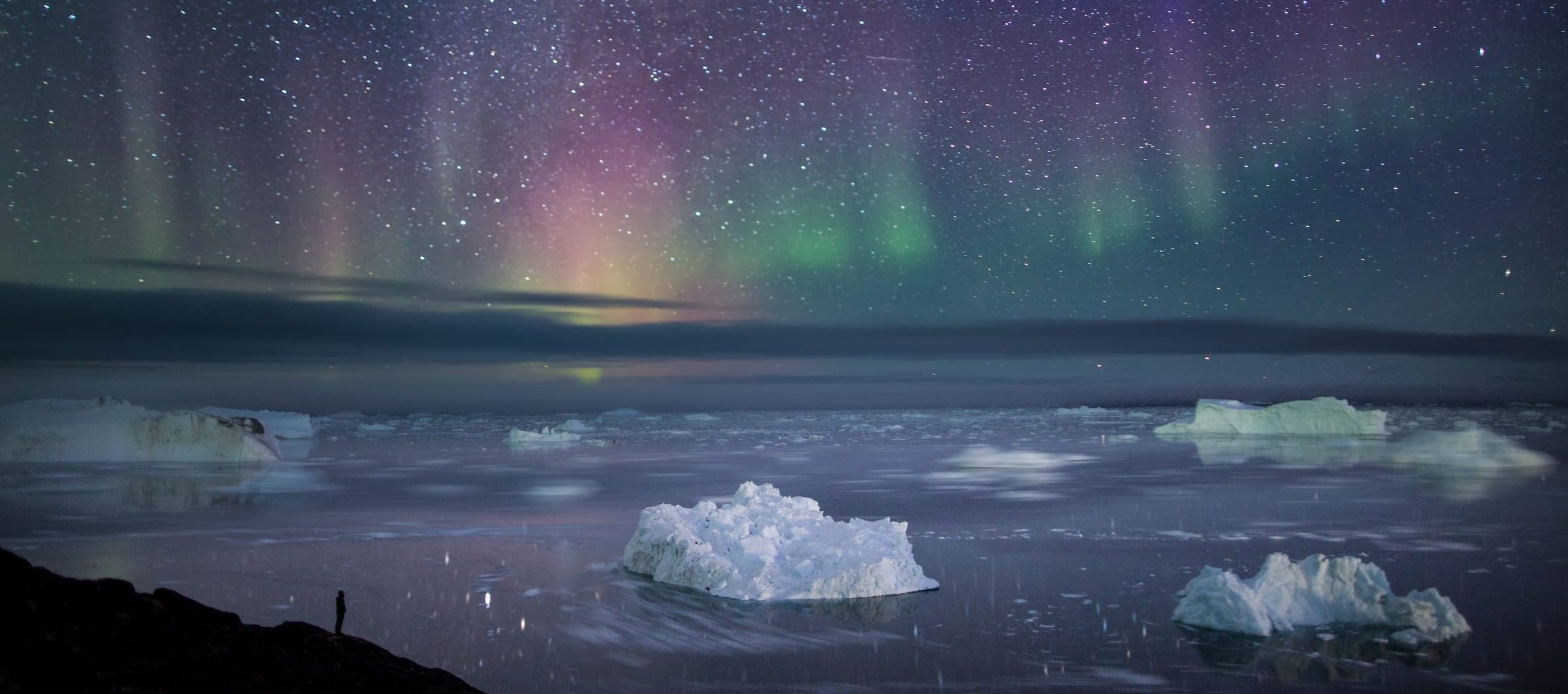 Hiker In Ilulissat, North Greenland Standing At The Sermermiut Overlook Close To The Ilulissat Icefjord Gazing At Icebergs And A Crisp Autumn Night Sky Filled With Stars And Northern Lights